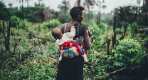 Kobieta odwrócona tyłem, na plecach niesie dziecko. Przed nią widać dużo roślinności