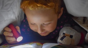 dziecko leży na łóżku i pod kołdrą z latarką w ręku czyta książkę