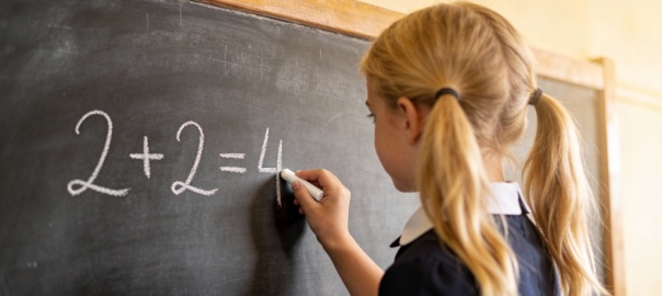 Dziewczynka stojąca przy tablicy szkolnej i zapisująca na niej zadanie z matematyki: 2+2=4  | fot. wygenerowany przez AI